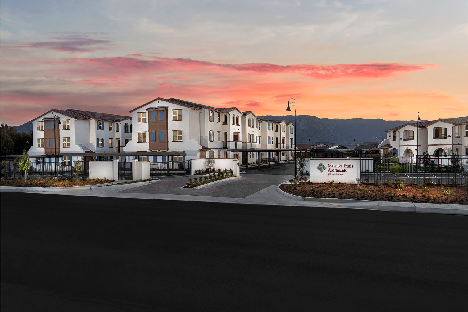 Mission Trail Apartments in Lake Elsinore: gated three-story buildings at sunset with mountains behind